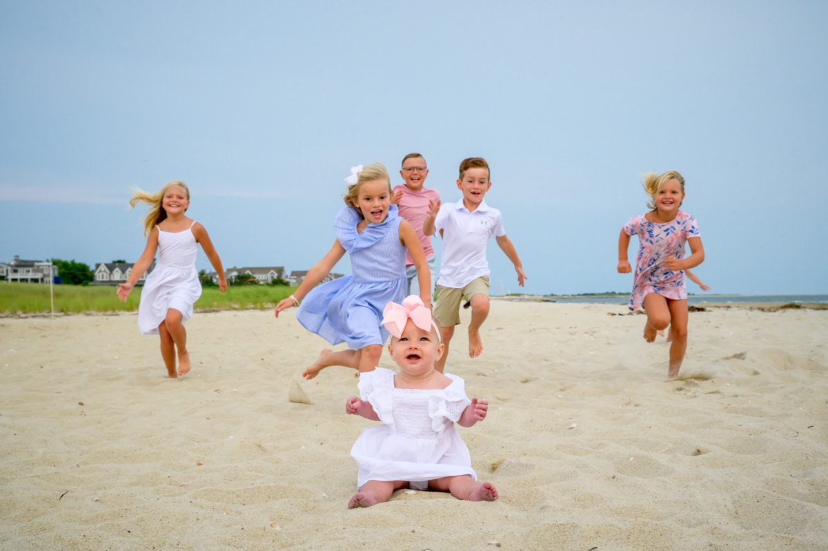 family-pictures-cape-cod-15 baby sitting on a beach in chatham, cape cod, with kids running towards her in the background